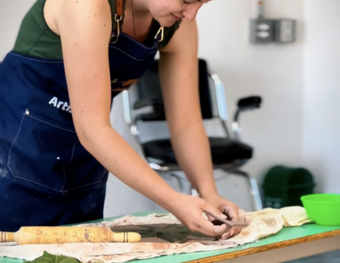 Mujer trabajando de forma artesanal sobre una mesa, manipulando materiales naturales con las manos y herramientas, en un espacio interior iluminado.