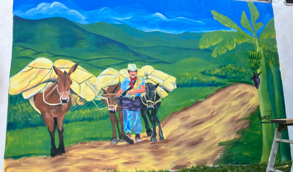 Mural colorido que representa a un arriero caminando por un camino rural acompañado de mulas cargadas, con montañas verdes y una mata de plátano al fondo.